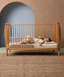 A toddler is lying in a cot with a low-profile pillow featuring safari animal print patterns. She is wearing a matching sleep suit bag and holding a plush lion.