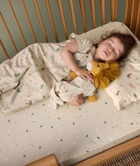 A toddler is peacefully asleep on a low-profile pillow with a safari-themed pillowcase. She is wearing a matching sleep suit bag and holding a plush lion.