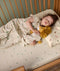 A young child is peacefully sleeping in a wooden toddler bed, wearing a safari-themed sleep suit bag and cuddling a plush lion toy dressed in a matching doll-sized sleeping bag.
