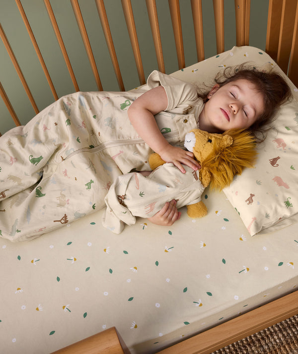 A young child is peacefully sleeping in a wooden toddler bed, wearing a safari-themed sleep suit bag and cuddling a plush lion toy dressed in a matching doll-sized sleeping bag.