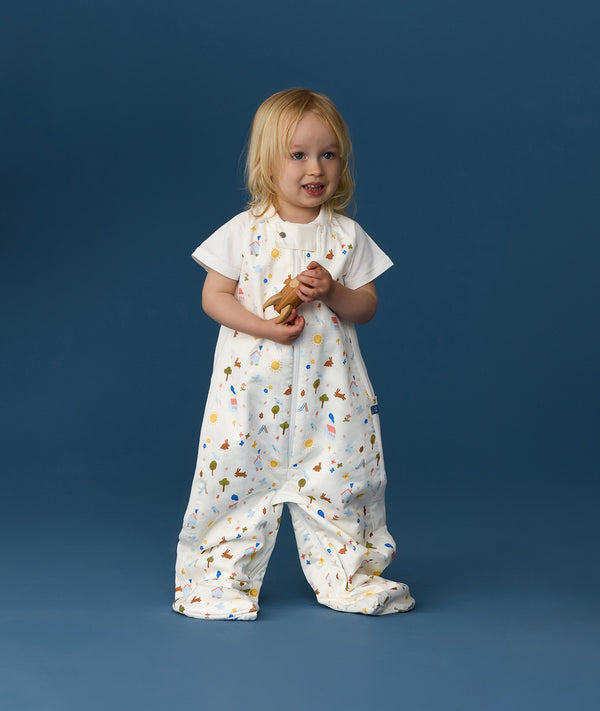 Toddler standing against a blue background wearing a white Sleep Suit Bag 1.0 TOG Daydream, featuring short sleeves and colourful all-over print