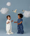 Two babies standing on a light blue background wearing 1.0 TOG Sleep Suit Bags, one in white with a colourful print and the other in blue with a moon-and-star print, reaching toward each other beneath decorative clouds and birds.