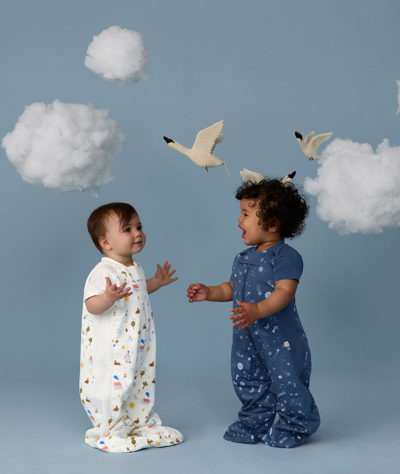 Two babies standing on a light blue background wearing 1.0 TOG Sleep Suit Bags, one in white with a colourful print and the other in blue with a moon-and-star print, reaching toward each other beneath decorative clouds and birds.