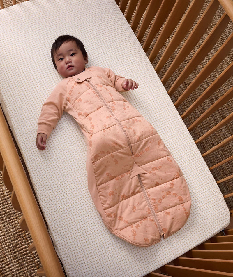 Baby lying in a wooden cot, wearing a peach-colored padded sleep suit bag with a front zipper and subtle animal print details.