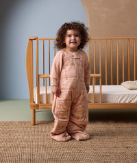 Smiling toddler standing in front of a wooden cot, wearing a peach-colored sleep suit with feet and a front zipper, decorated with a subtle animal pattern, in a cozy nursery setting.