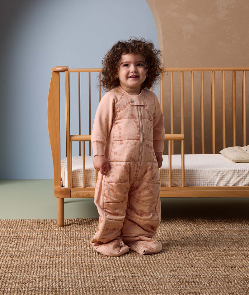 Smiling toddler standing in front of a wooden cot, wearing a peach-colored sleep suit with feet and a front zipper, decorated with a subtle animal pattern, in a cozy nursery setting.