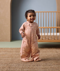 Smiling toddler standing in front of a wooden cot, wearing a peach-colored sleep suit with feet and a front zipper, decorated with a subtle animal pattern, in a cozy nursery setting.