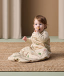An infant sits on a rug, wearing an ergoPouch Sleep Suit Bag, and puts a toy in its mouth.