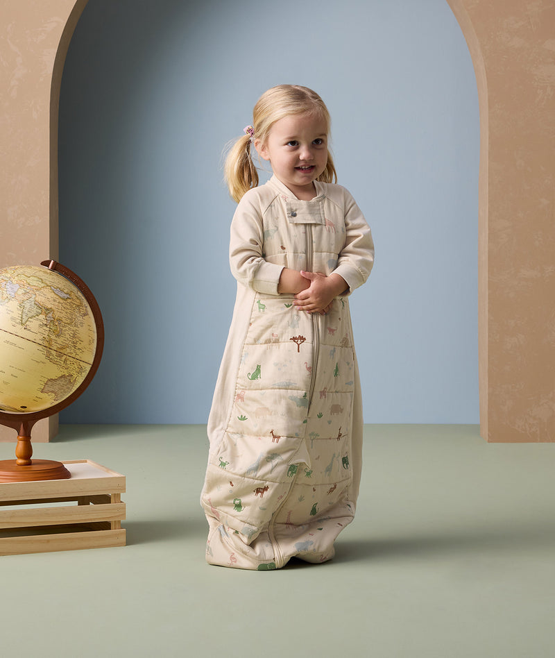 Smiling toddler standing indoors wearing a beige sleep suit bag with colorful safari animal prints, next to a globe placed on a small wooden stand.