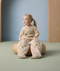 Toddler sitting on a woven cushion, wearing a beige sleep suit bag with legs, featuring a colorful animal print pattern including giraffes, elephants, and other safari animals.