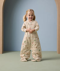 Young child standing and smiling indoors, dressed in a beige sleep suit bag with legs with colorful safari animal prints, against a soft blue and beige arched background.
