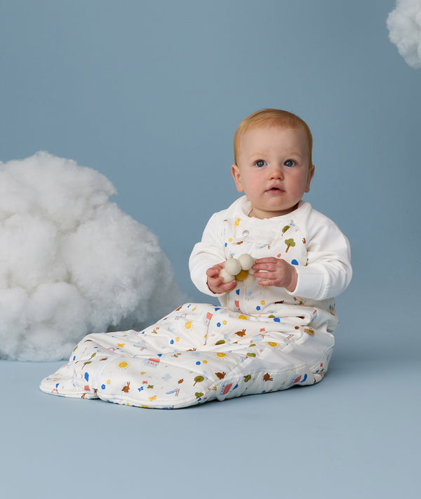 Infant sitting upright against a light blue background with decorative clouds wearing a white Sleep Suit Bag with sleeves