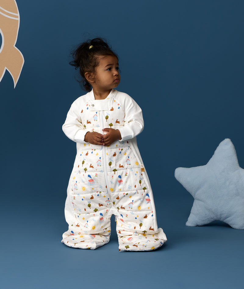 Toddler standing against a blue background wearing a Sleep Suit Bag 2.5 TOG Daydream, featuring a white base with colourful all-over print.