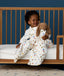 Toddler sitting on a bed wearing a Sleep Suit Bag 2.5 TOG Daydream while a holding a plush toy dressed in a matching sleeping bag
