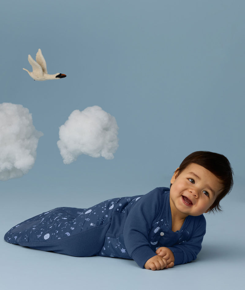 Infant crawling against a light blue background wearing a 2.5 TOG Sleep Suit Bag, featuring a stretchy side panel for natural movement.