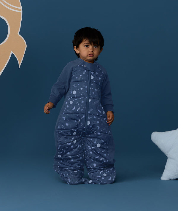 Toddler standing against a deep blue background wearing a Sleep Suit Bag 2.5 TOG Starbeam, featuring long sleeves and star and moon print.