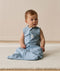 Infant sitting on a white mat, wearing a blue sleeveless Sleeping Bag with front zipper.