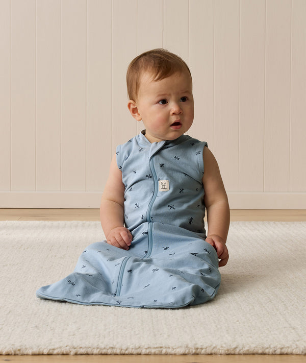 Infant sitting on a white mat, wearing a blue sleeveless Sleeping Bag with front zipper.