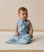 Infant sitting on a white mat, wearing a blue sleeveless Sleeping Bag with front zipper.