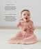 A baby sitting on a cream rug, holding a wooden ring toy to its mouth while wearing a pink floral sleeping bag with text highlighting organic cotton and stretchy & bell-shaped design.