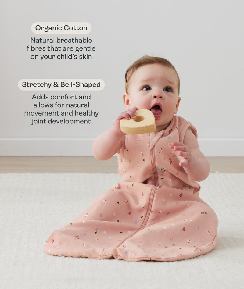A baby sitting on a cream rug, holding a wooden ring toy to its mouth while wearing a pink floral sleeping bag with text highlighting organic cotton and stretchy & bell-shaped design.
