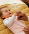 A close-up view of a baby lying on a mustard-yellow quilted mat while an adult gently adjusts the zipper of the baby's pink floral sleeping bag with text overlay highlighting slim-fit design for an easy transition from swaddling.
