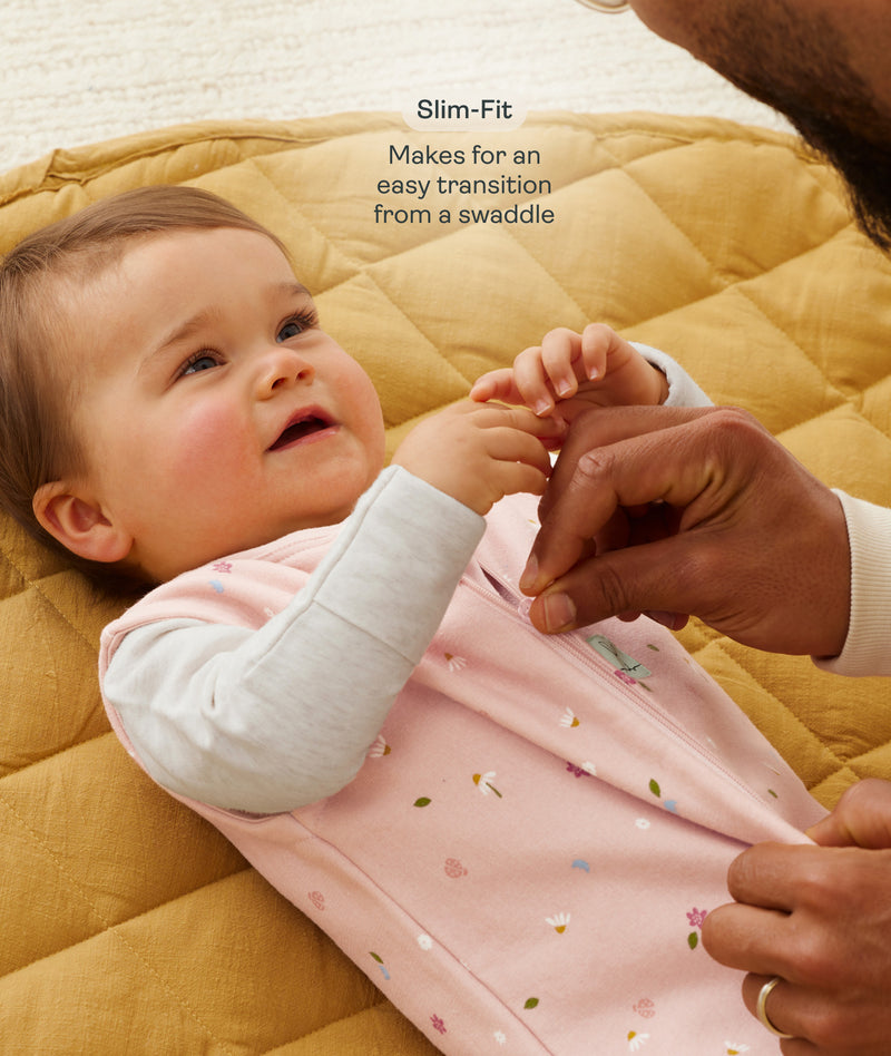 A close-up view of a baby lying on a mustard-yellow quilted mat while an adult gently adjusts the zipper of the baby's pink floral sleeping bag with text overlay highlighting slim-fit design for an easy transition from swaddling.