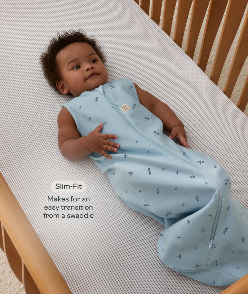 Infant lying on their back inside a wooden cot wearing a sleeveless dragonfly-patterned Sleeping Bag with text highlighting slim-fit design for  an easy transition from swaddling.