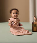 The infant is sitting on the floor holding a crocheted toy, wearing an ergoPouch Sleeping Bag with elephant print.