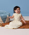 A happy baby sitting on a brown mat wearing a sleeveless, cream sleeping bag with a subtle tan pattern and front zipper.