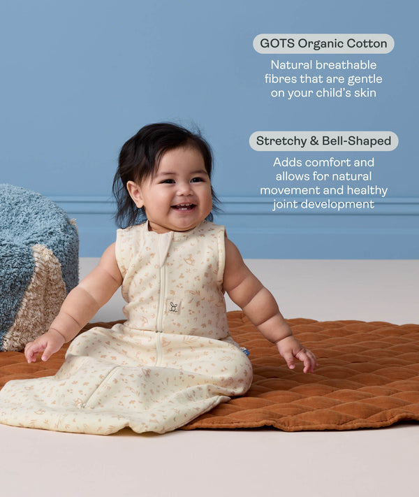 A happy baby sitting on a brown mat wearing a sleeveless, cream sleeping bag with text highlighting 