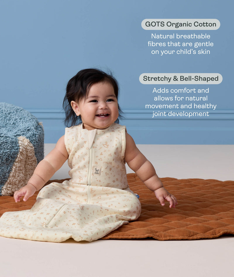 A happy baby sitting on a brown mat wearing a sleeveless, cream sleeping bag with text highlighting 