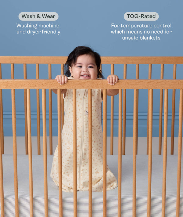 A smiling baby standing in a wooden cot wearing a cream-colored, pattern-printed sleeping bag with text highlighting 