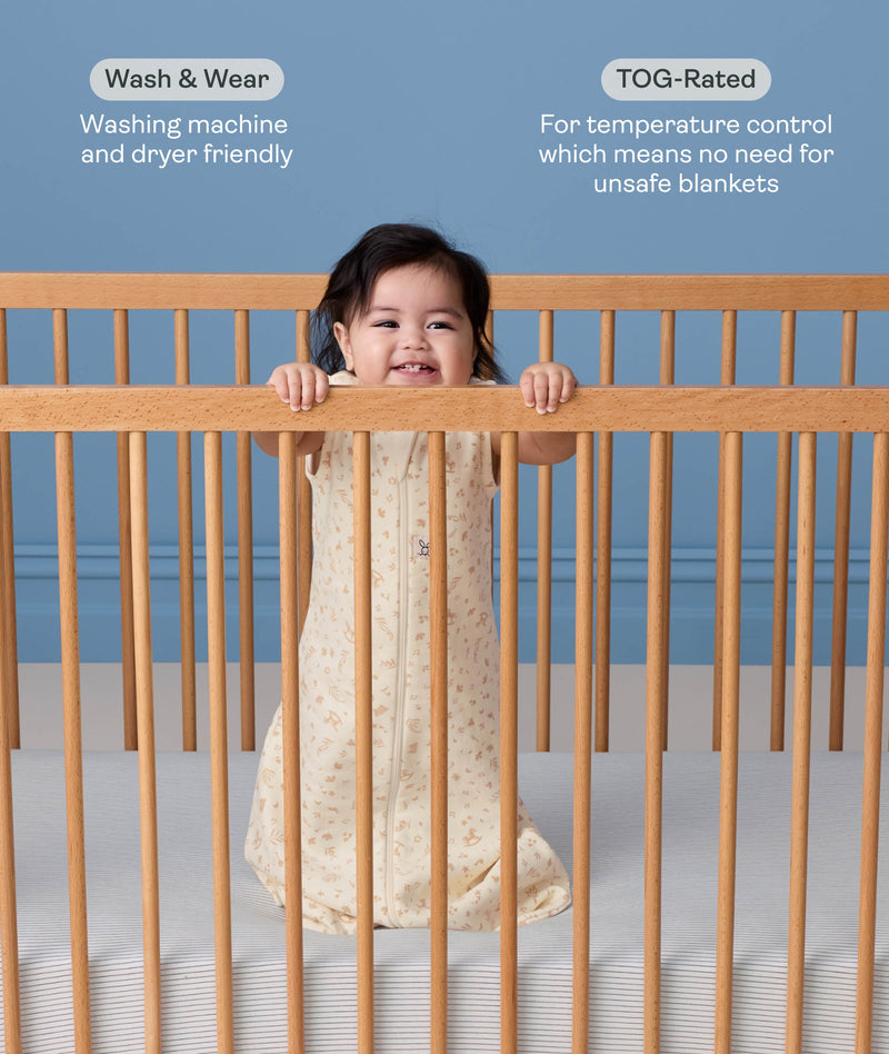 A smiling baby standing in a wooden cot wearing a cream-colored, pattern-printed sleeping bag with text highlighting 