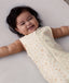 A happy baby lying on a pinstriped crib sheet, wearing a sleeveless cream sleeping bag with a subtle tan pattern and a front zipper.