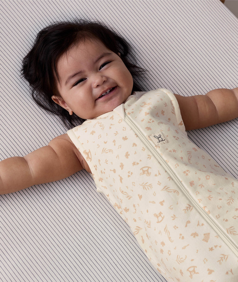 A happy baby lying on a pinstriped crib sheet, wearing a sleeveless cream sleeping bag with a subtle tan pattern and a front zipper.