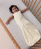 A happy baby lying in a wooden cot, wearing a sleeveless cream sleeping bag with a subtle tan pattern and a full-length zipper.