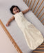 A happy baby lying in a wooden cot, wearing a sleeveless cream sleeping bag with a subtle tan pattern and a full-length zipper.