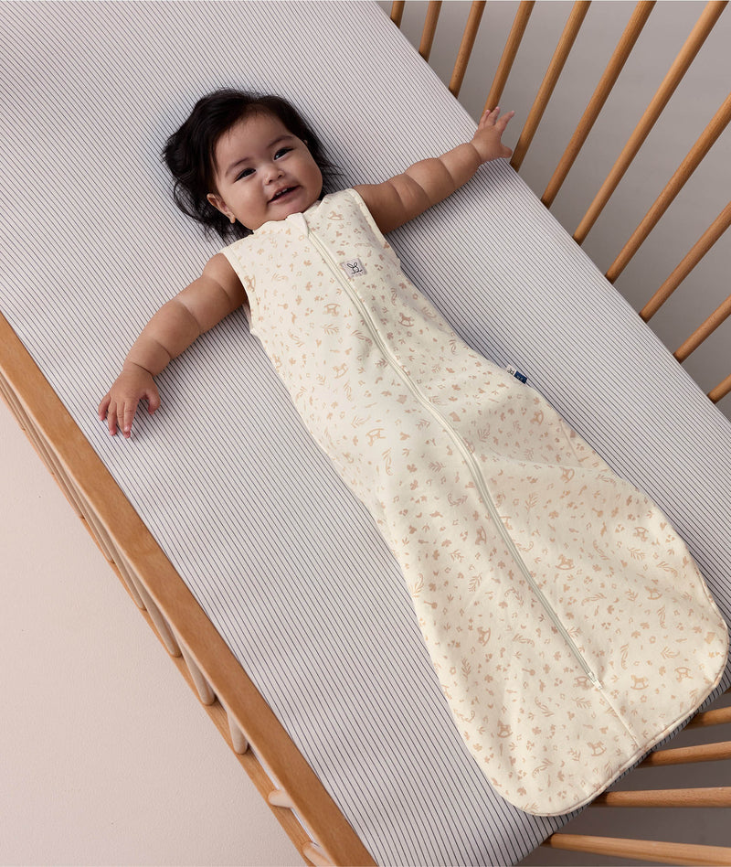 A happy baby lying in a wooden cot, wearing a sleeveless cream sleeping bag with a subtle tan pattern and a full-length zipper.