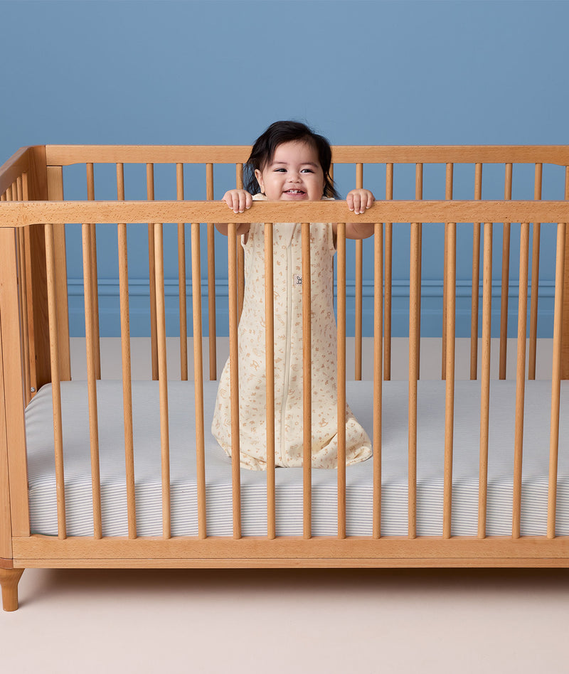 A smiling baby standing in a wooden cot wearing a cream-colored, pattern-printed sleeping bag.