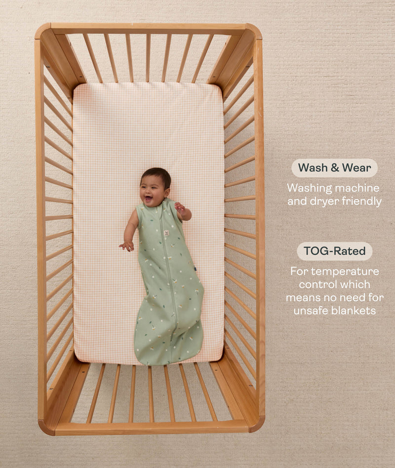 A happy baby dressed in a sleeveless ergoPouch Sleeping Bag 1.0 TOG, lying in a wooden cot with text highlighting 