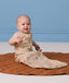Infant sitting on a brown quilted rug, wearing an ergoPouch Sleeping Bag while holding a wooden toy on one hand.