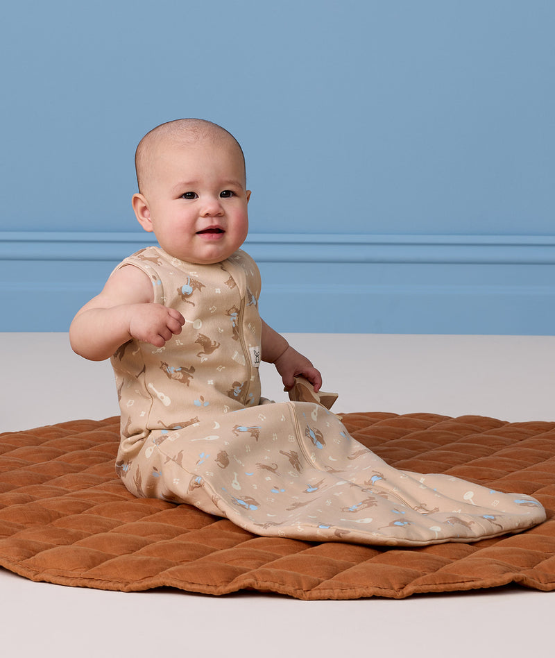 Infant sitting on a brown quilted rug, wearing an ergoPouch Sleeping Bag while holding a wooden toy on one hand.