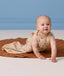 A smiling baby crawling on a round brown mat, wearing a sleeveless beige baby sleeping bag featuring a light blue and brown patterned print.