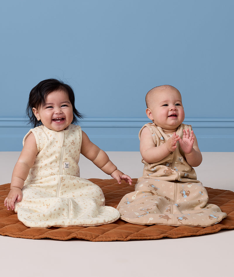 Two happy babies sitting on a mat wearing sleeveless, pattern-printed sleeping bags.