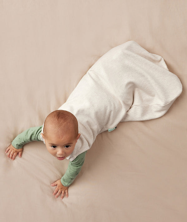 Baby crawling on a plain surface while wearing an oatmeal-coloured sleeping bag over a green long sleeved layer.