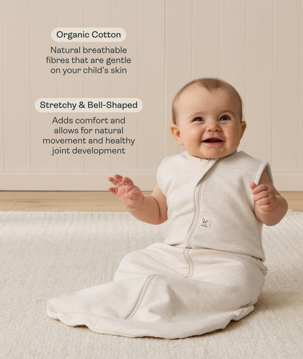 Smiling infant sitting on a white mat, wearing a sleeveless Sleeping Bag in Oatmeal Marle, with text highlighting organic cotton and stretchy, bell-shaped design.
