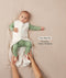 A baby lying down in a green long sleeve layer with an unzipped oatmeal-coloured sleeping bag while adult hands hold their feet. Text highlighting 