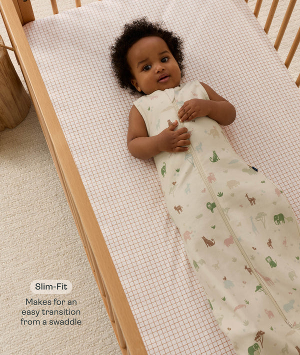 An overhead view of a smiling baby lying in a wooden cot wearing an animal-patterned Sleeping Bag, featuring a 