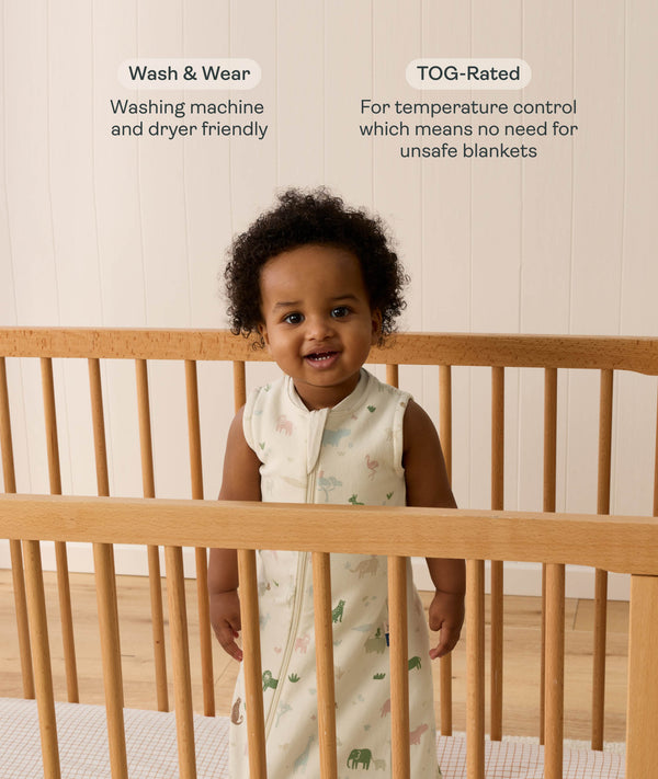 A toddler standing inside a light wooden cot wearing an animal-patterned 1.0 TOG Sleeping Bag, accompanied by product feature callouts for 